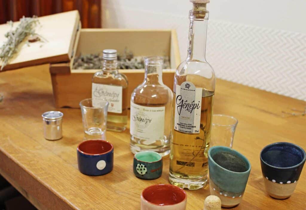 Une sélection de trois bouteilles de Génépi présentées avec des branches séchées de génépi dans une boîte et des tasses de dégustation en céramique