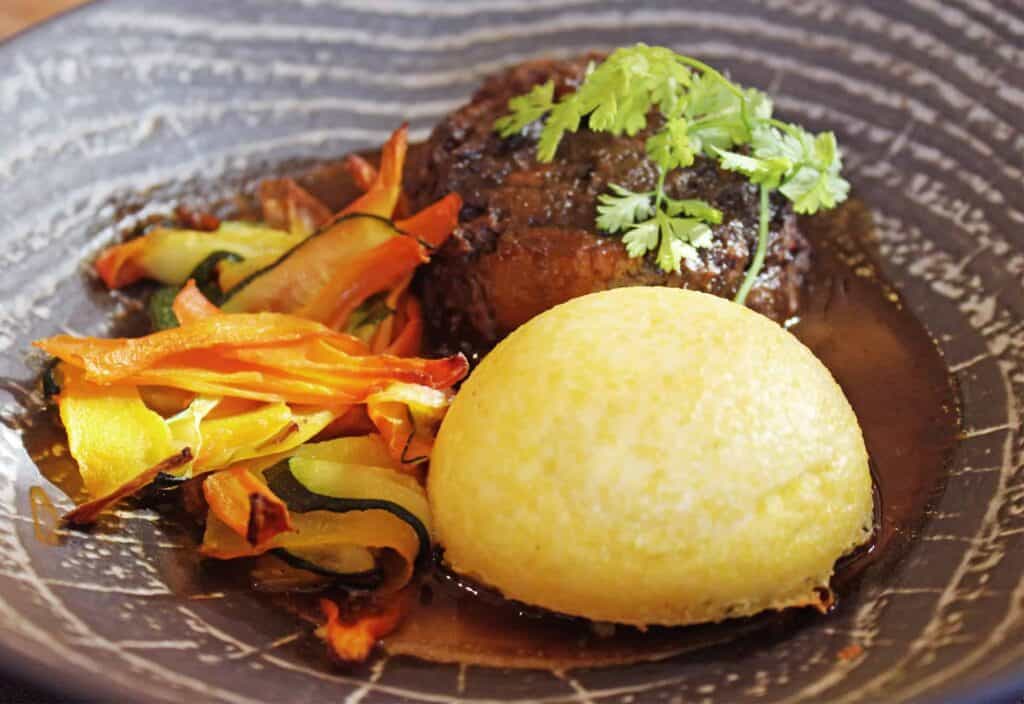 A beef dish with polenta and roasted vegetables in gravy on a black plate