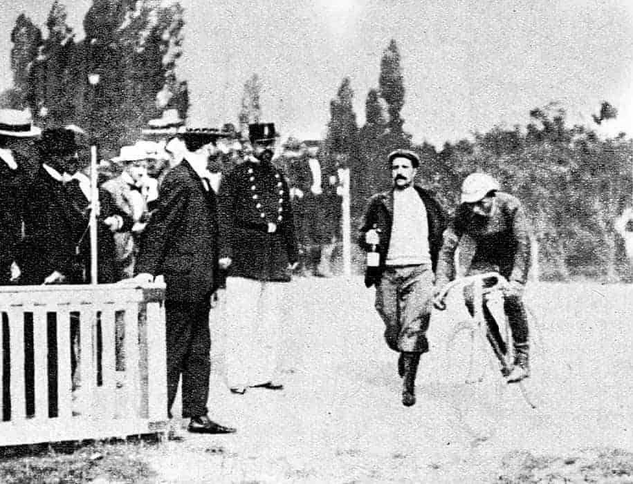 Maurice Garin cycling for the finish line with a man running behind him