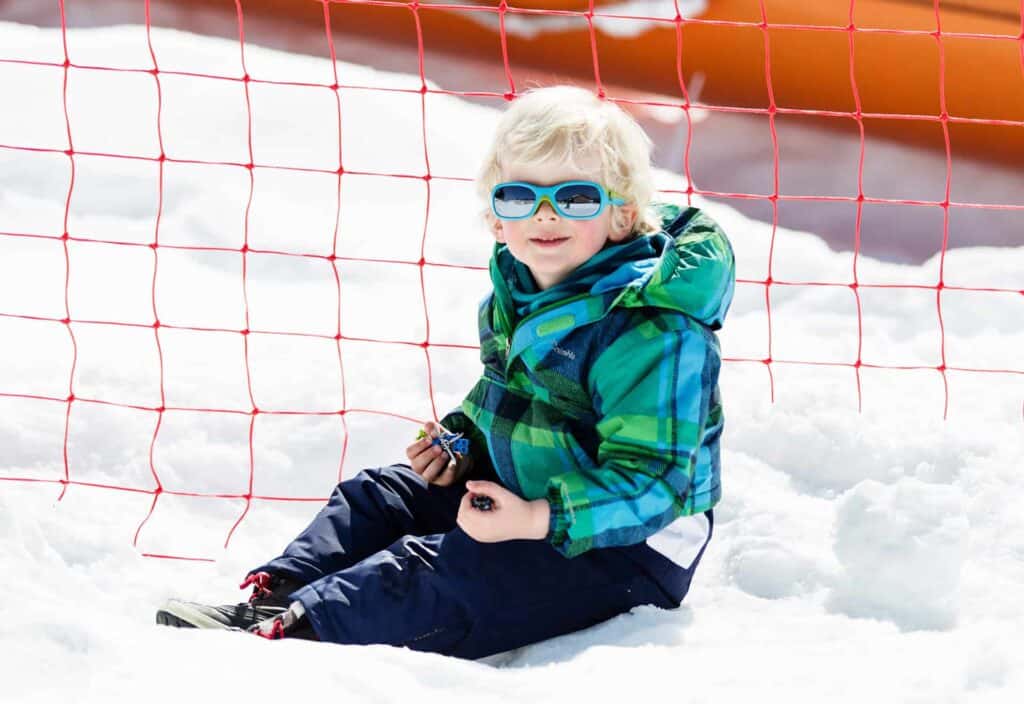 A young blond boy sat in the snow wearing green and blue ski wear and green sunglasses