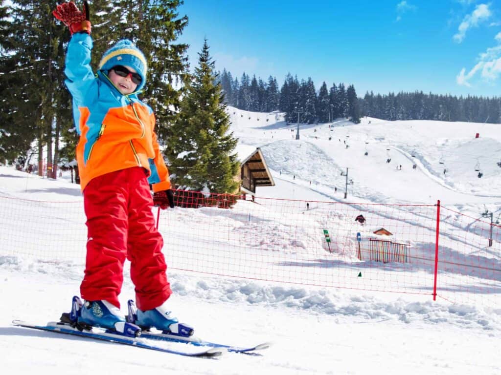 A happy child dressed in colourful ski clothes skiing on a sunny ski slope during their winter family holiday.