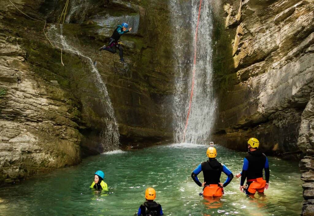 A group canyoning under a large waterfall and one man is jumping from the rocks into the pool