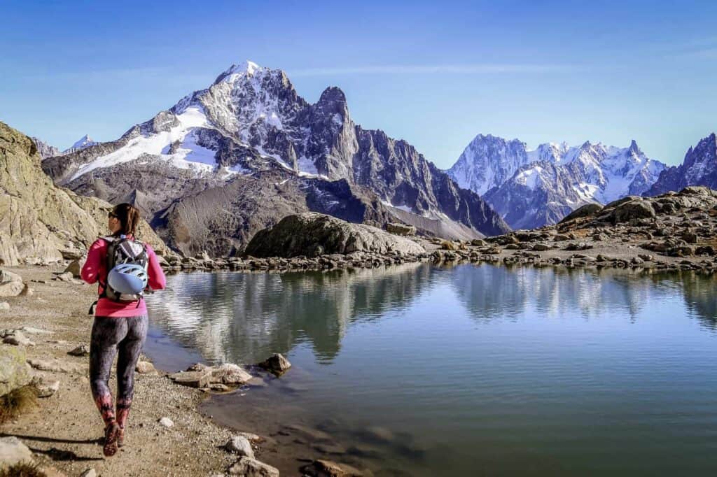 A woman hikes along a lake, with snowy mountains in the background.