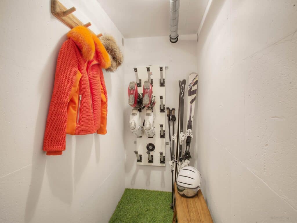 A white ski room with an orange ski jacket hanging up and a boot and ski rack in the background