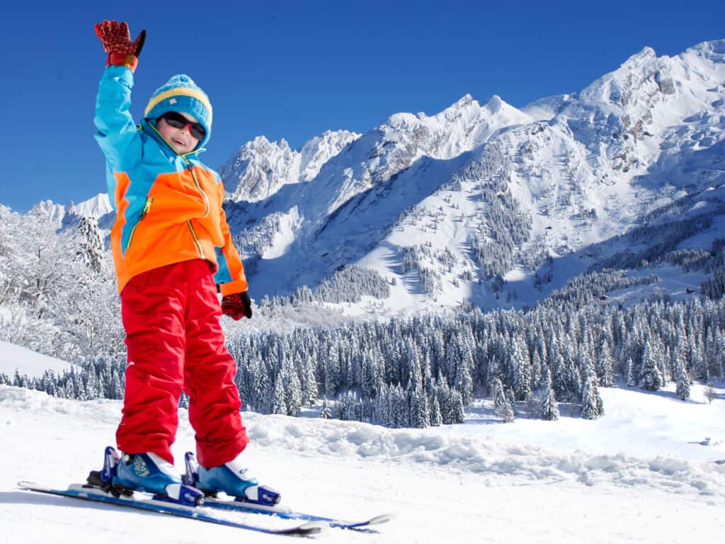 Young child skiing and waving with mountains in the background.