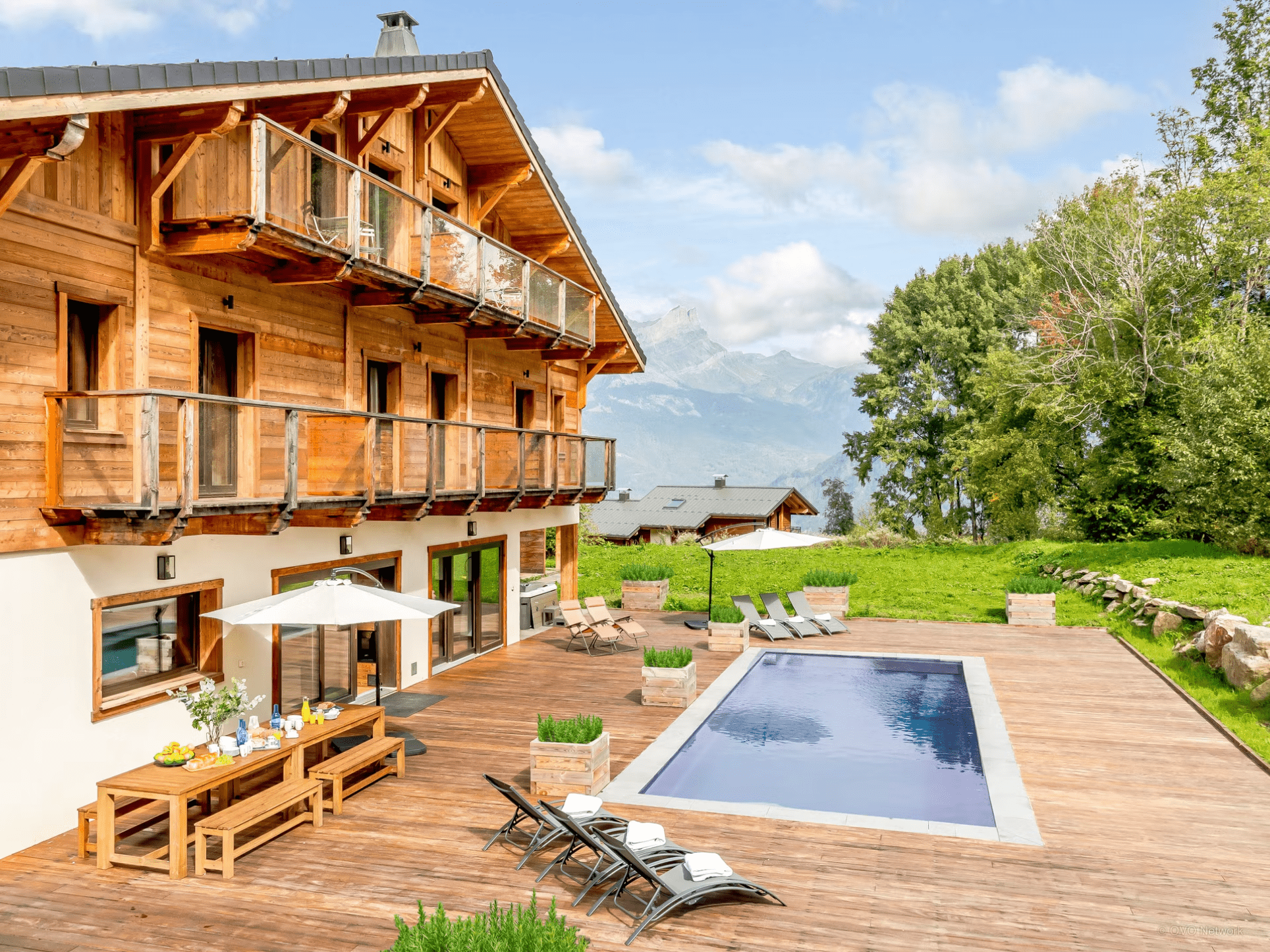 Extérieur d'un chalet en bois à plusieurs étages dans les Alpes. Une piscine se trouve à l'extérieur avec chaises longues, parasol et jardin. 