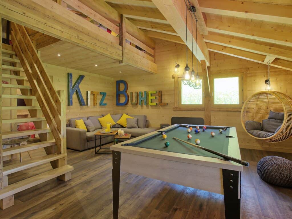 Large text art spelling 'Kitz Buhel' hangs on a wooden wall in a playroom with a pool table and sofas.