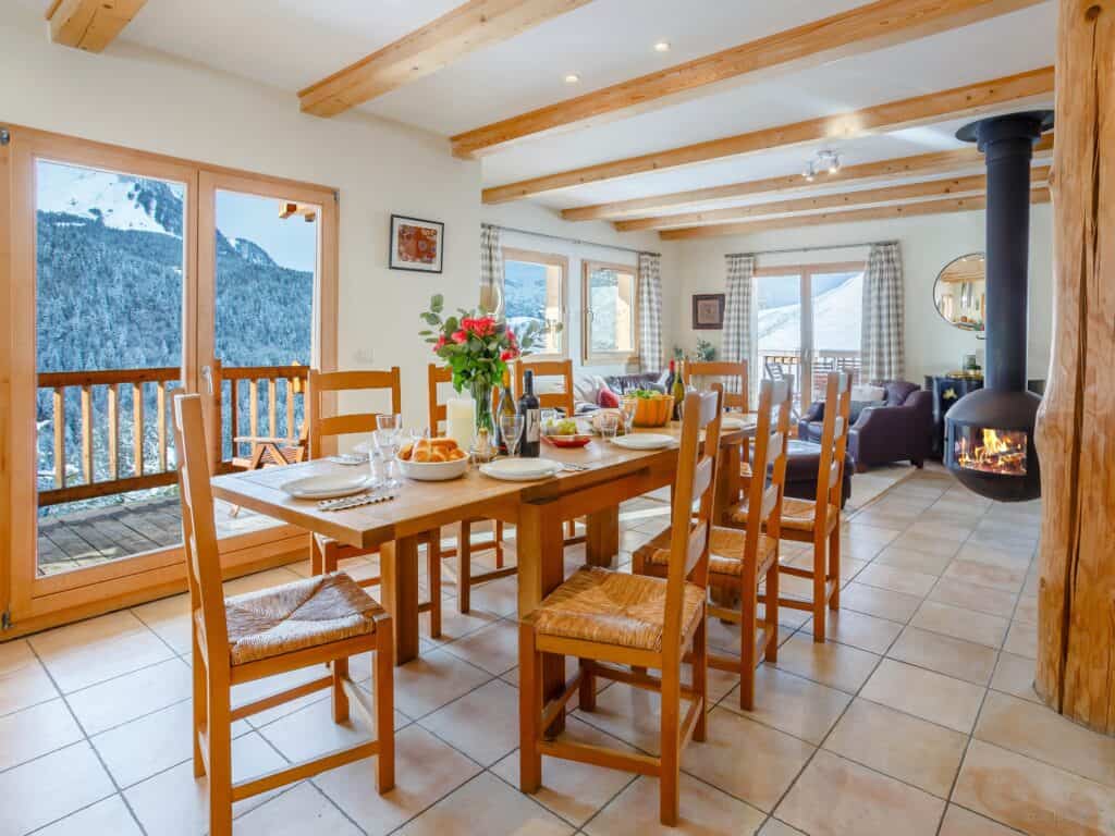 Vaste salle à manger avec vue sur la montagne. Carrelage clair au sol, table et chaises en bois, poêle à bois et larges fenêtres constituent le décor.