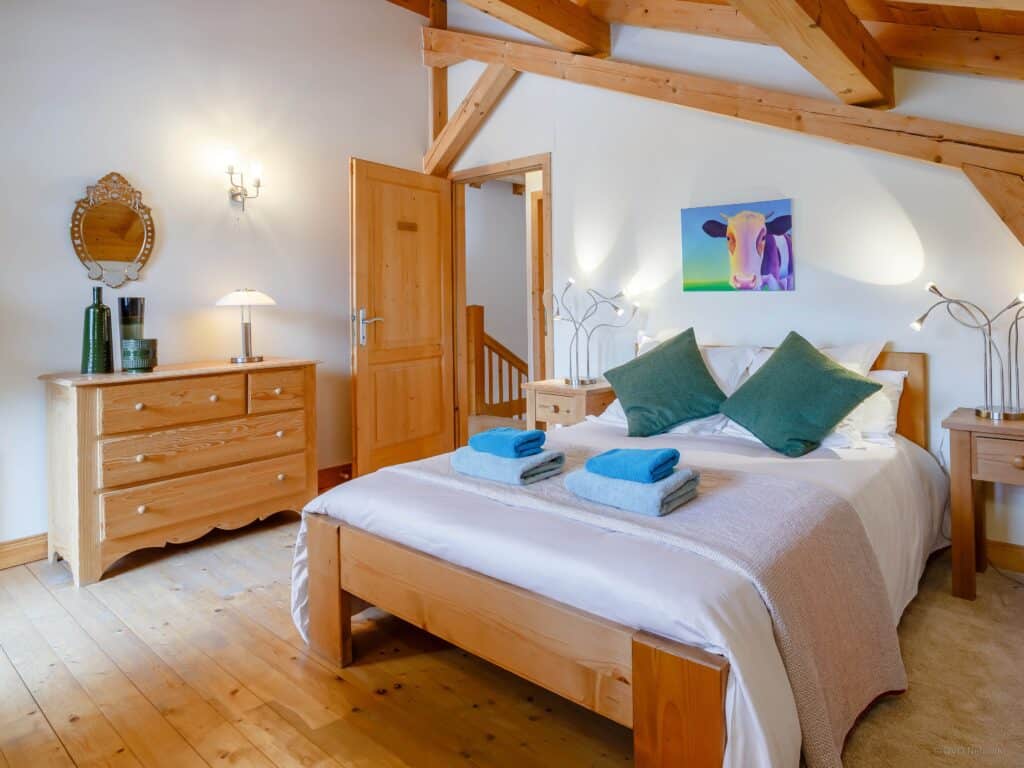 Chambre double avec coussins colorés et linge de lit clair. Au mur se trouve un tableau représentant une vache.  