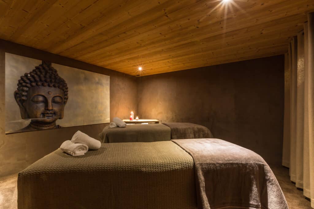 Une pièce aux couleurs sombres avec deux tables de massages, des serviettes pliées et un tableau représentant Bouddha. 
