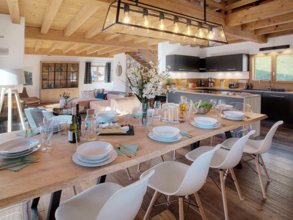 Espace de vie avec table à manger entièrement dressée, chaises claires et luminaire allumé. Parquet au sol et plafond en bois avec poutres. Au fond se trouve la cuisine.