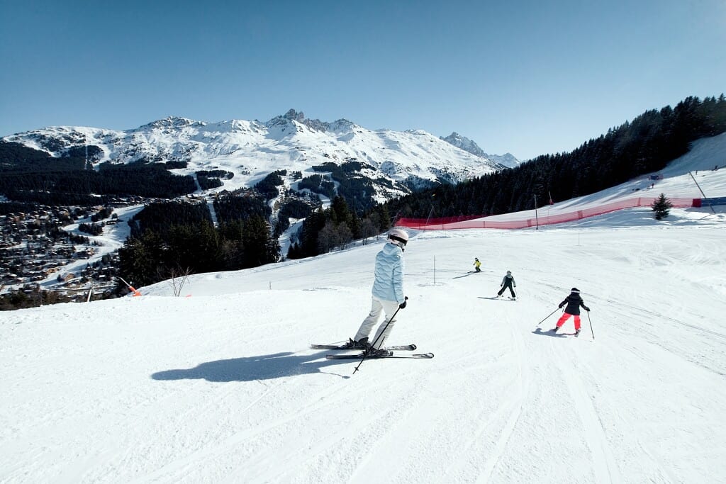 Four people ski down a piste in Meribel, with a village on the left and mountains and forests in the background