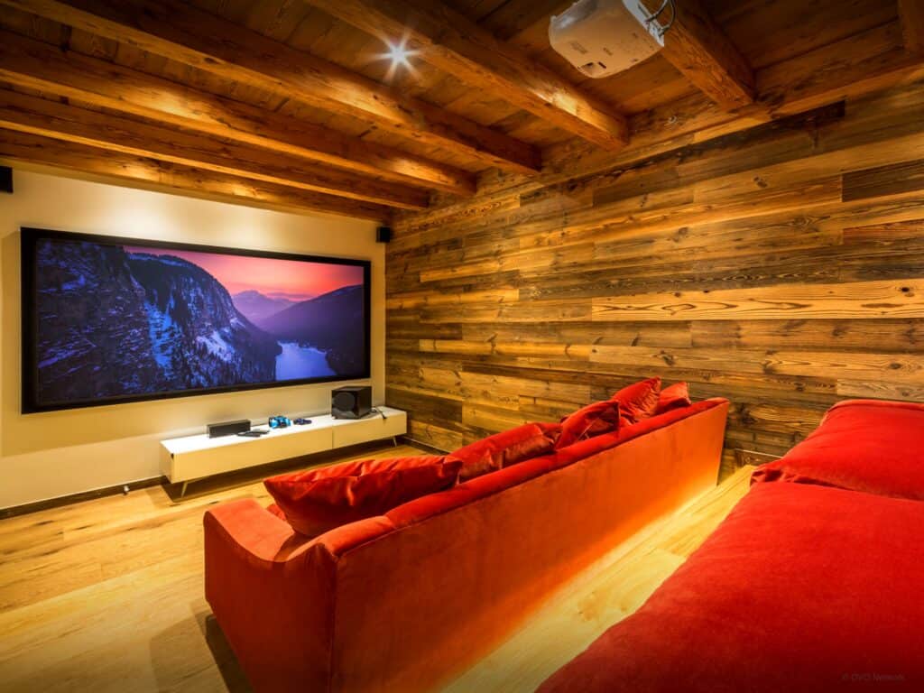 A cinema room with red sofas on two levels and a wide screen, with natural wood walls