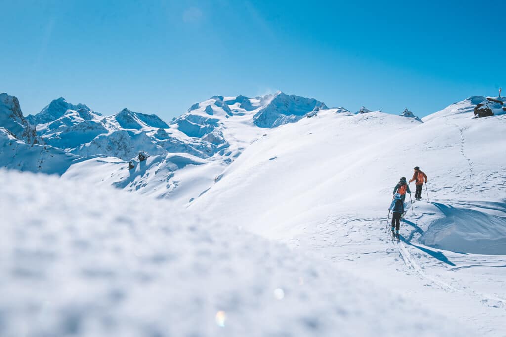 A blue sky over snowy mountains as three people enjoy a ski-touring excursion