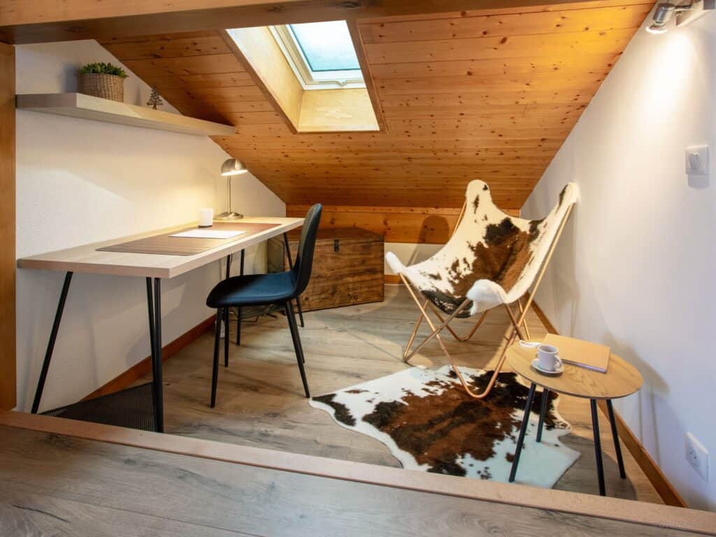 Un espace de travail à domicile avec une fenêtre velux, un bureau, une chaise imprimée vache et une lampe de bureau.