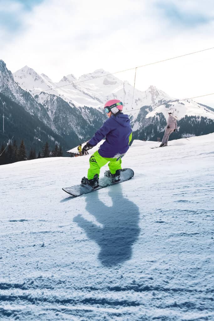 Person snowboarding in the Aravis area, wearing green trousers, blue coat, red helmet, and glasses. In the background, the mountains. 