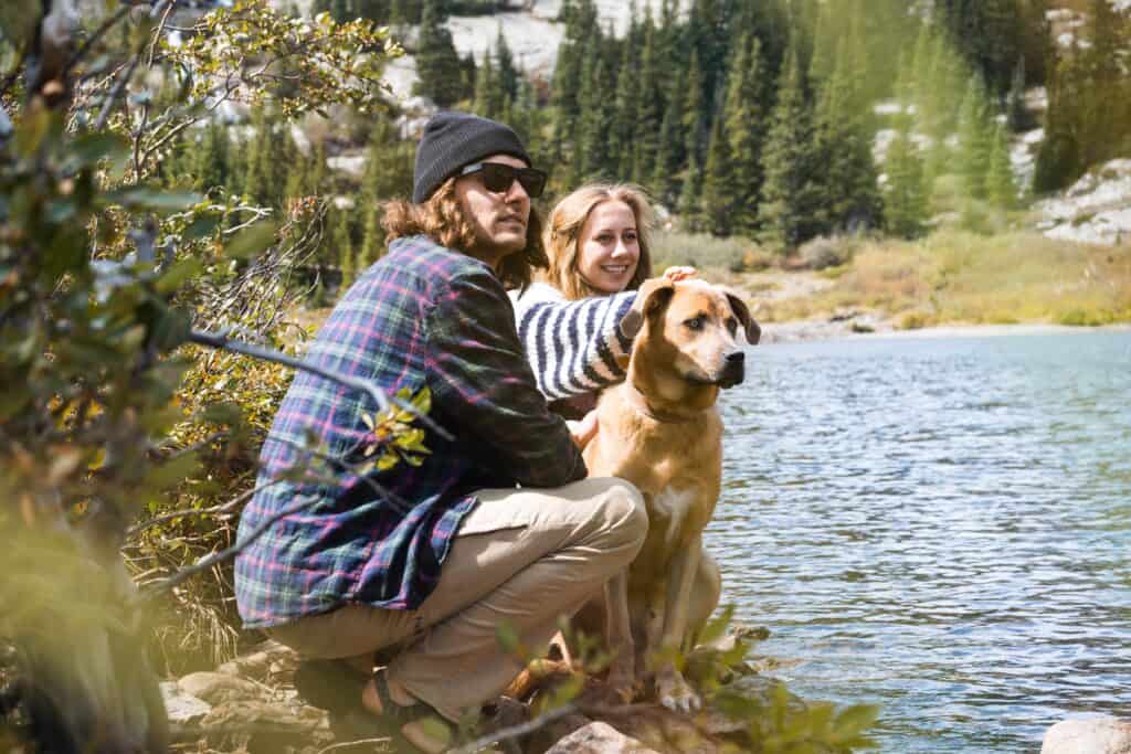 A couple sit by a lake with their dog. The man wears a checked shirt and sunglasses and the woman wears a striped sweater.