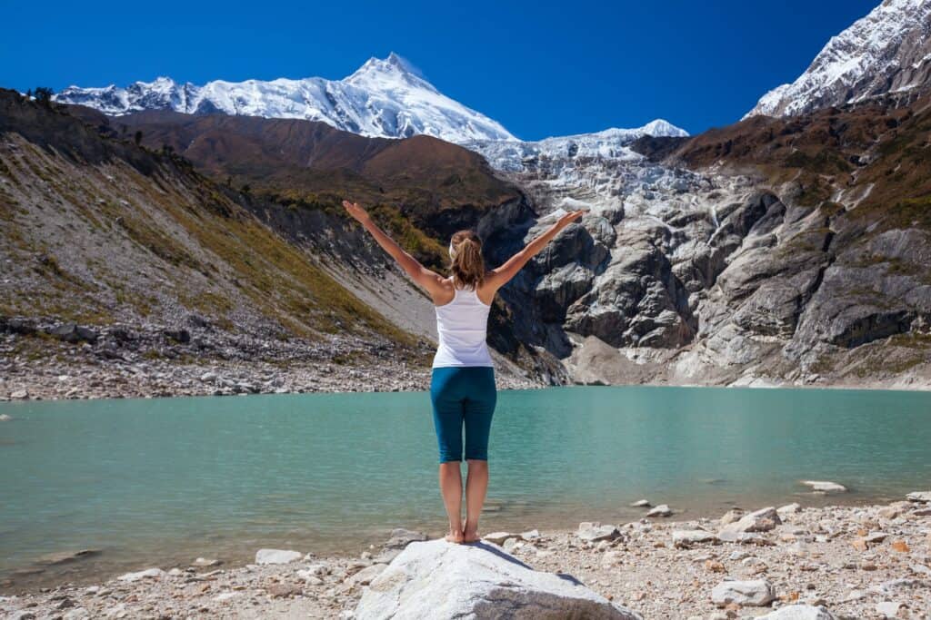 Une femme se tient debout, dos à la caméra, face à un lac de montagne, les bras levés vers le ciel.