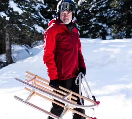 A man wearing a helmet stands in the snow with his wooden sledge