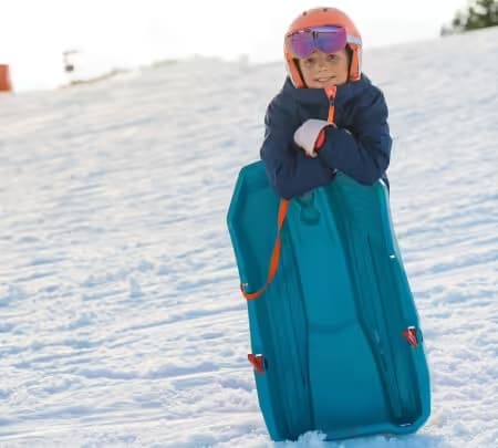 A child stands on the snow, leaning on their sledge