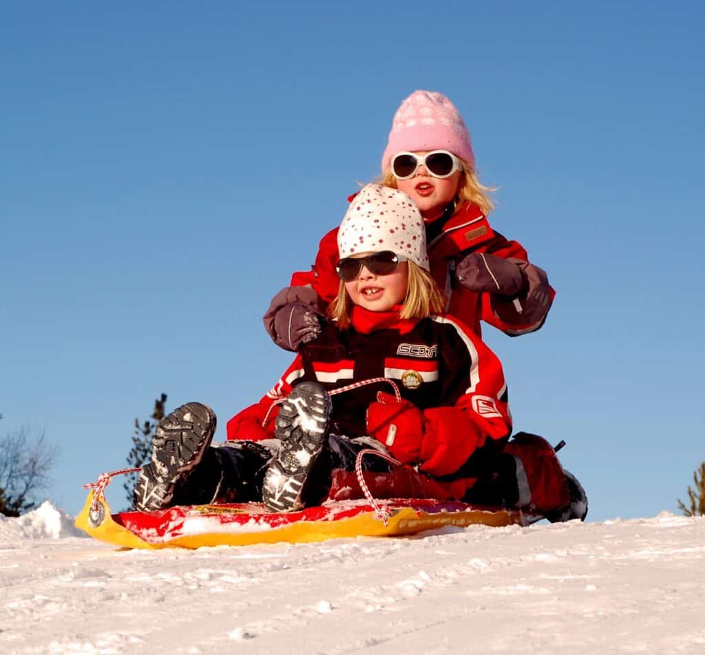 Two young children sledging in the mountains, wearing snowsuits, hats and sunglasses