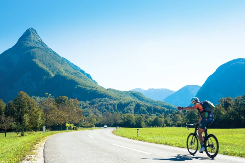 A man cycles along an Alpine road holding his thumb out