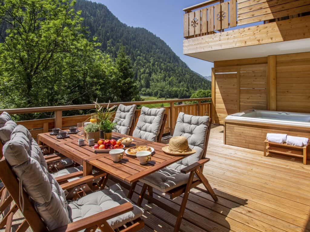 A sunny balcony with a dining table set for 6 and a hot tub overlooking the mountains