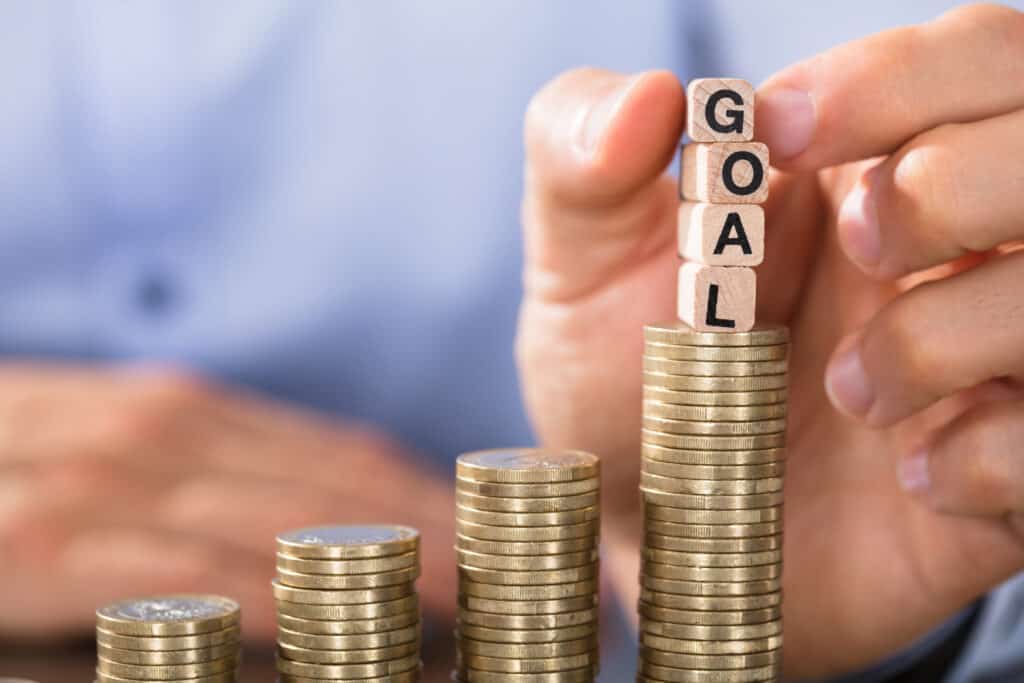 Close-up of a hand holding blocks spelling out the word GOAL over a large pile of coins