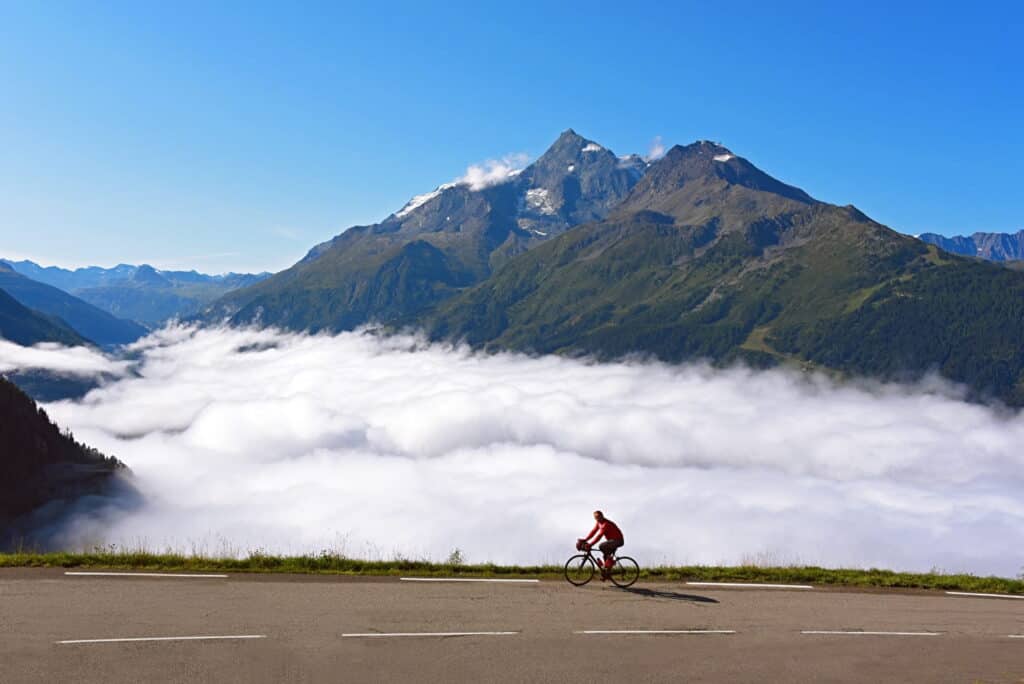 A cyclist on a road above cloud level, with mountain tops 
