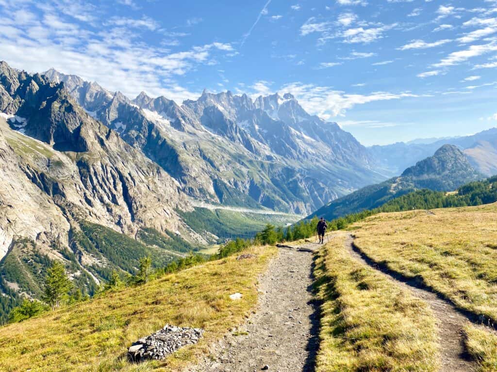 An image of a man trekking up a mountain path with large mountain peaks in the background in the Aosta Valley in Italy