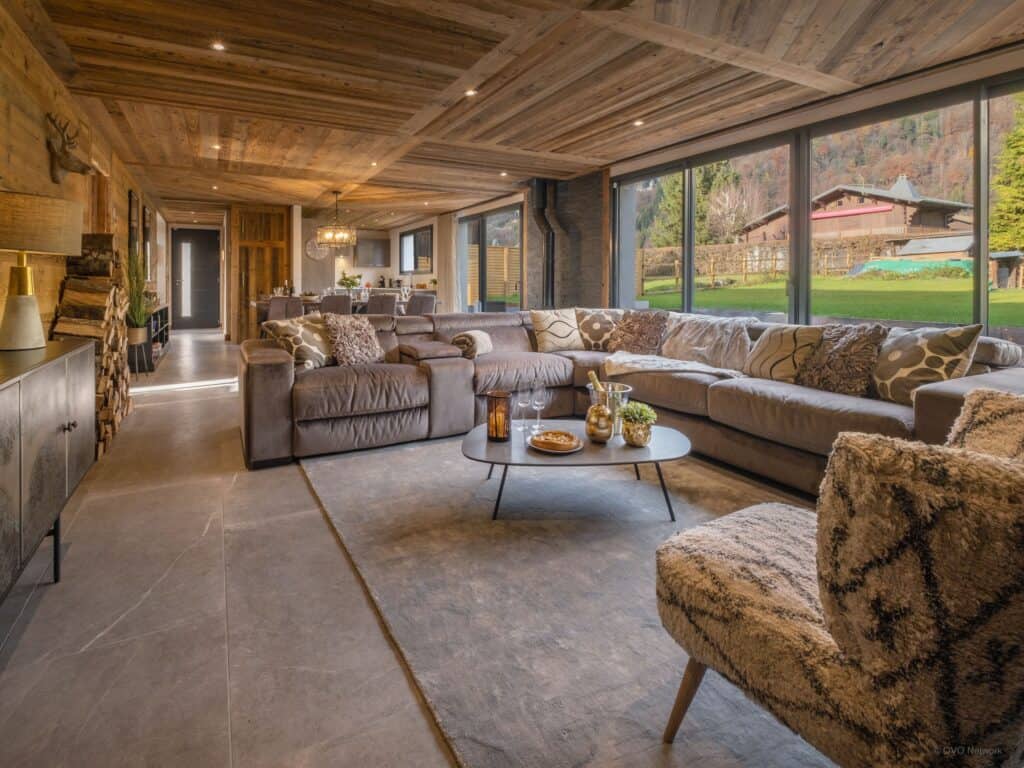Comfortable sofas and chairs in front of large windows in the liviing room of Chalet des Hottes in Morillon