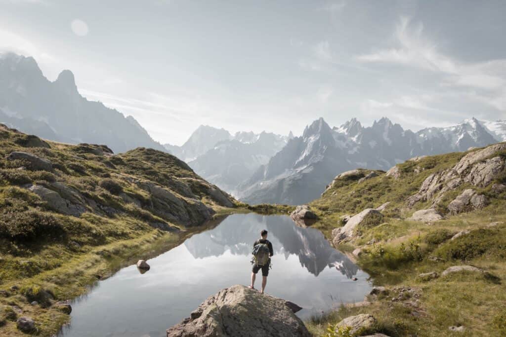 A hiker stopped on a lake shore, with mountains in the background.
