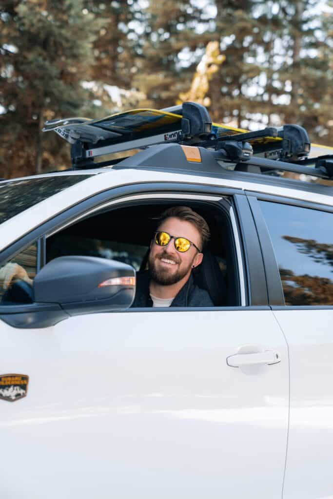 Un homme portant des lunettes de soleil regarde par la fenêtre d'une voiture transportant des skis et un snowboard.