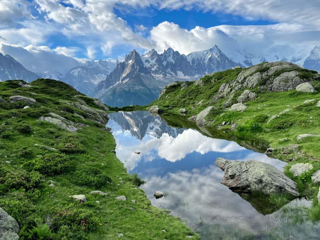 A clear mountain lake reflecting the jagged mountain peeks surrounded by green pastures and rocks