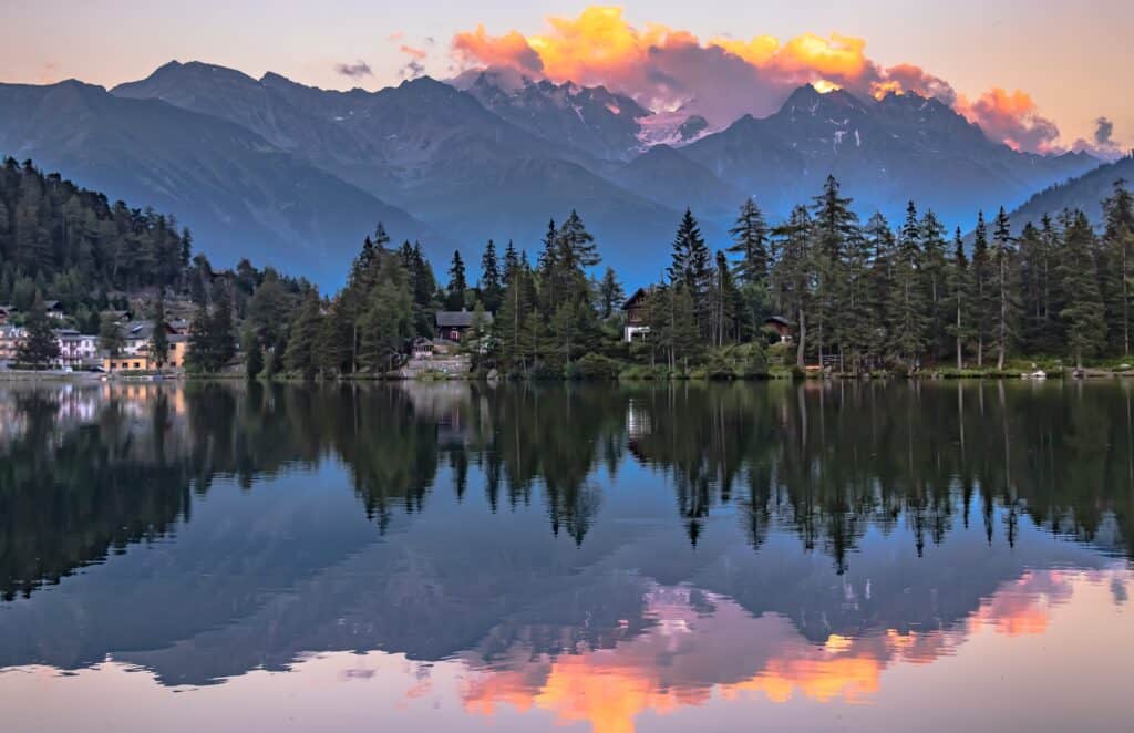 An Alpine lake at sunset, reflecting pink skies and clouds, surrounded by trees and lakeside huts