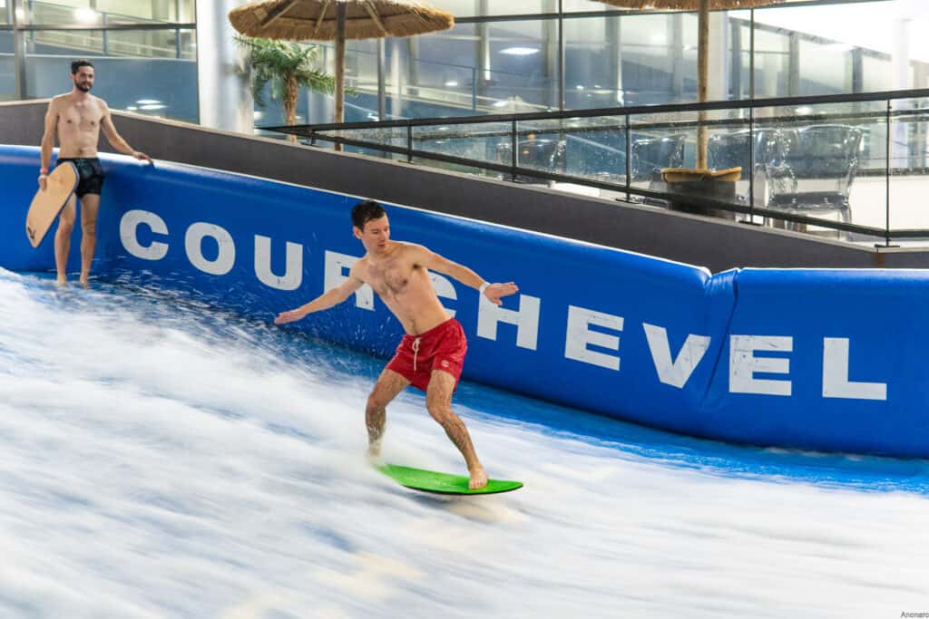A man wearing red swim shorts tries indoor surfing as his friend watches
