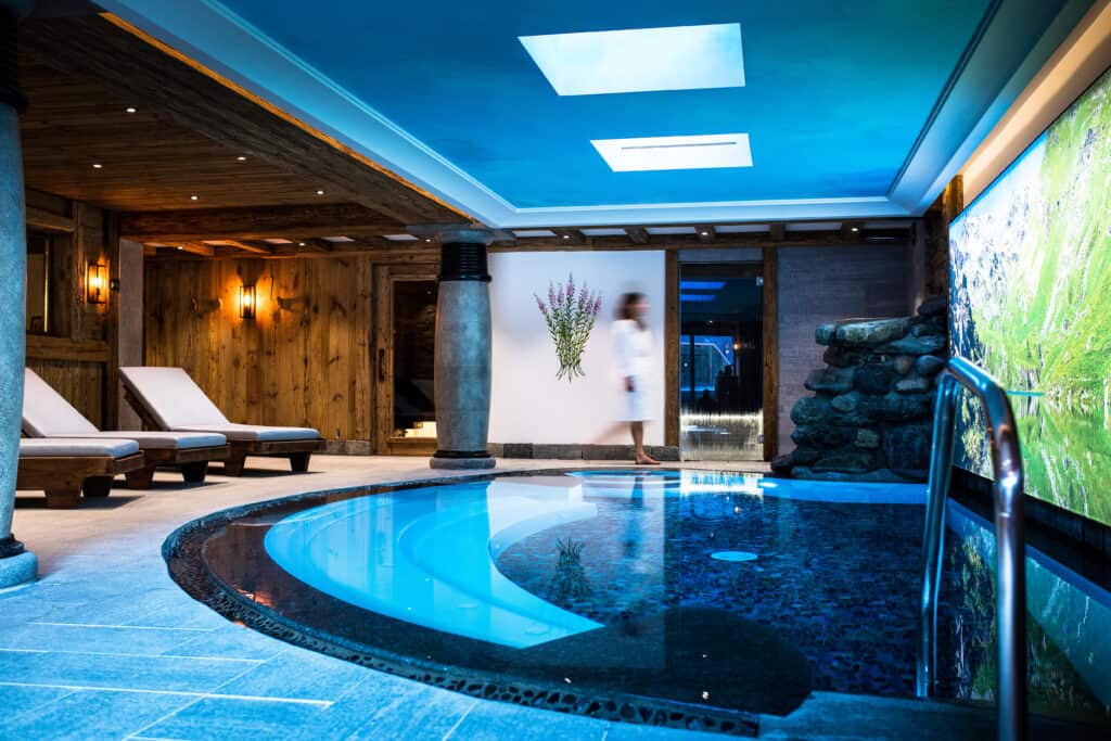 An indoor swimming pool with a large screen displaying images of the mountains and deck chairs around the edge