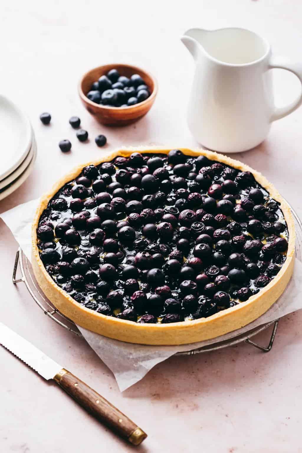 A bilberry tart on a cooling rack, with fresh bilberries and a jug of cream
