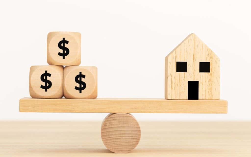 A wooden seesaw with money on one side and a house on the other