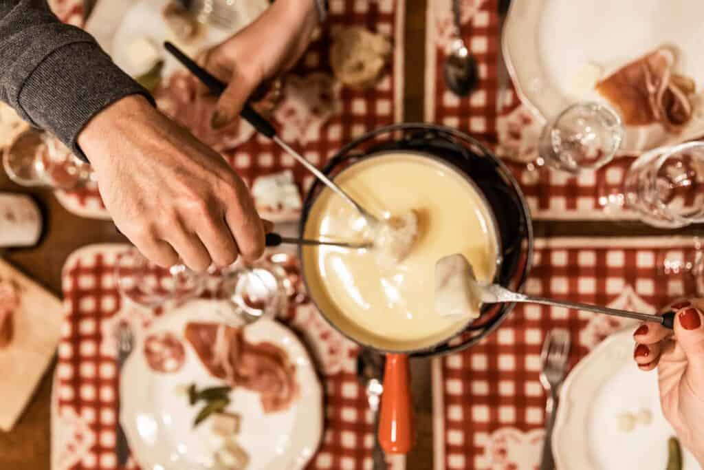 Three people dip their bread in a cheese fondue, surrounded by plates of cold meat and pickes