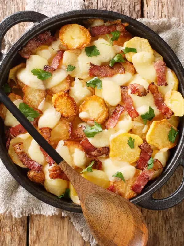A dish of potatoes, bacon, cheese and herbs laid out on a table with a wooden spoon.