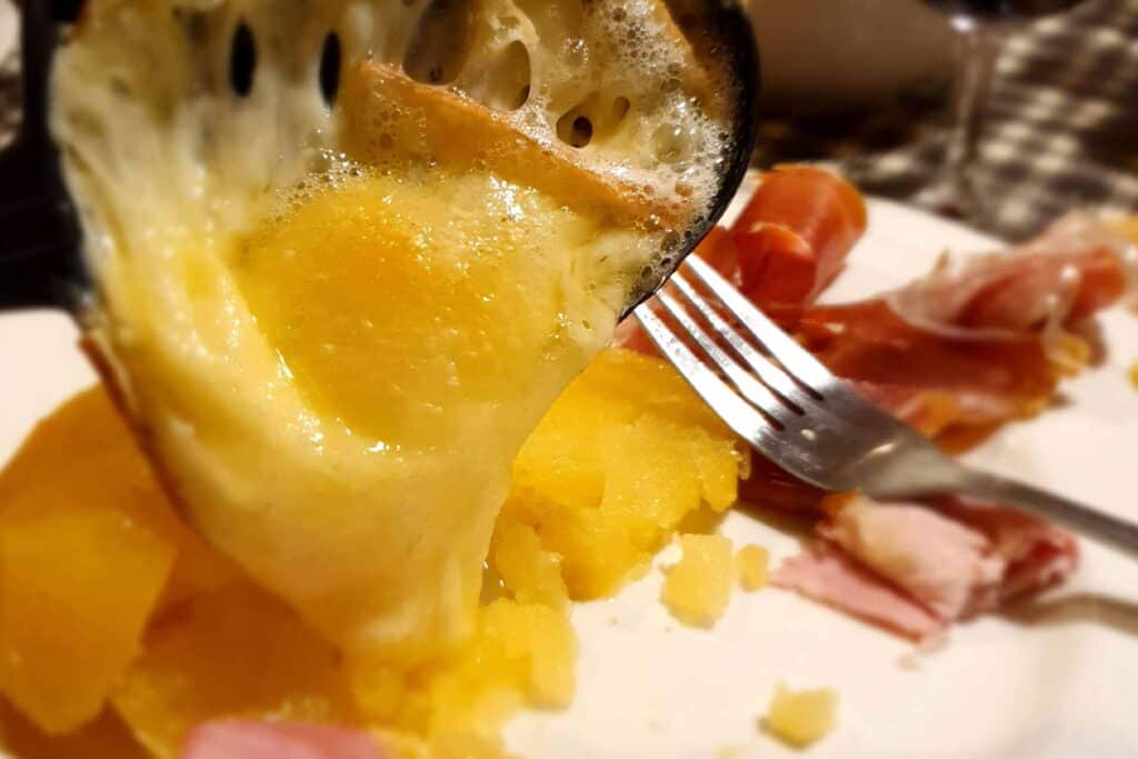 Melting raclette cheese is poured on to a white plate, with potatoes and cold meats