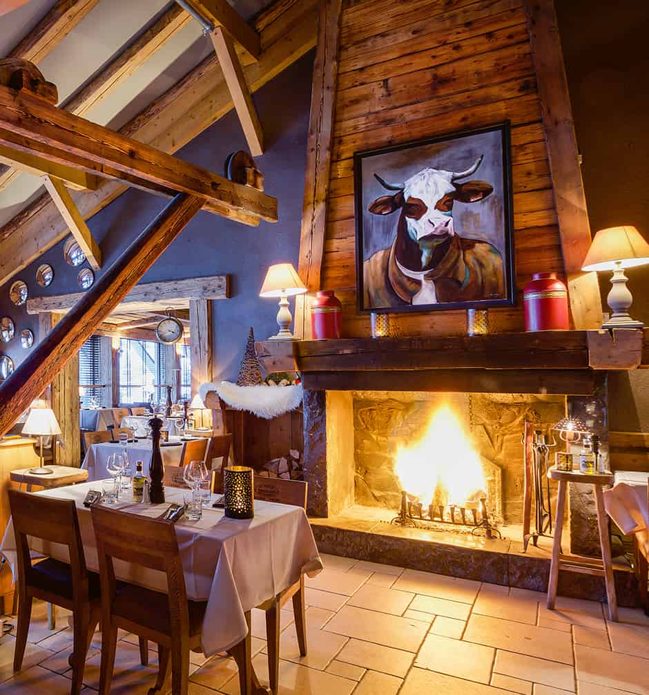Intérieur d'un restaurant avec un tableau de vache et une cheminée allumée, dans un chalet en bois.