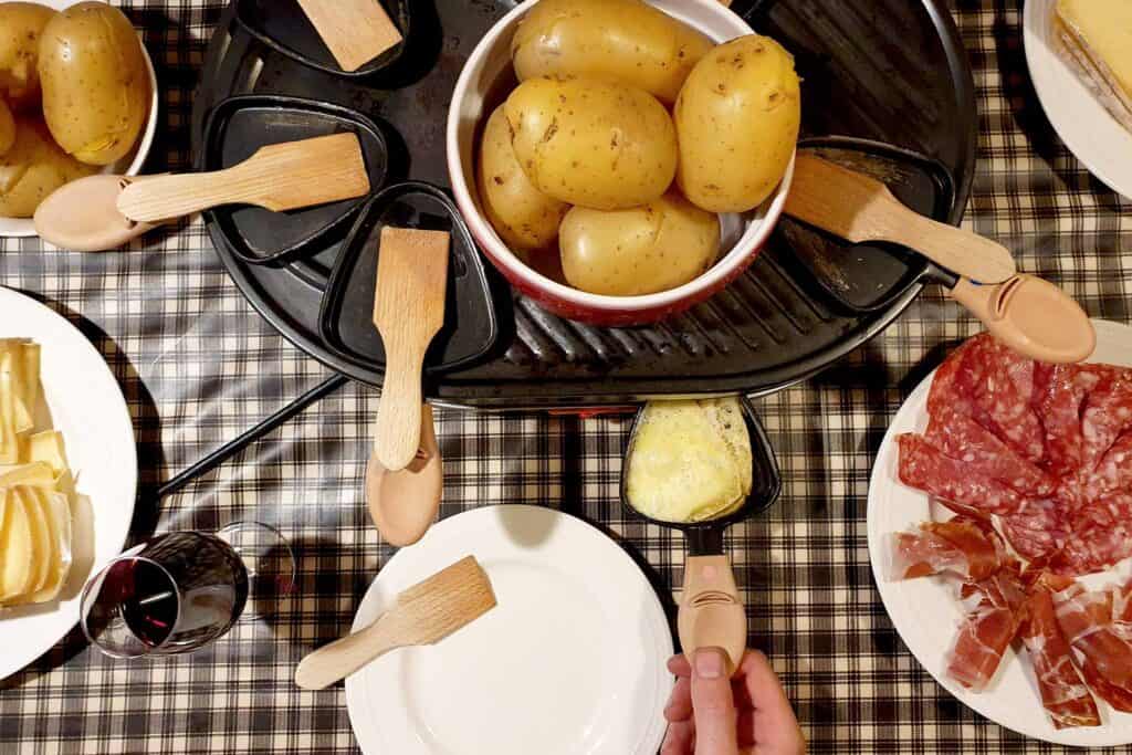 An electric raclette device on a checked tablecloth, with potatoes, charcuterie and red wine