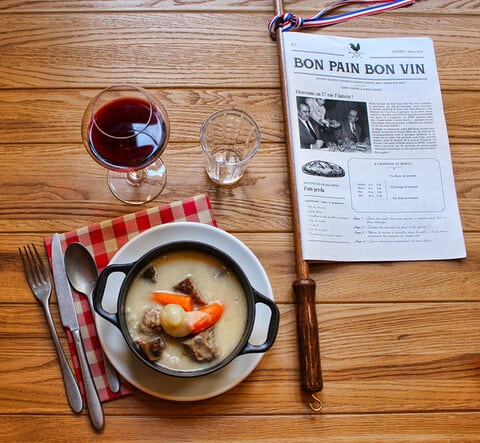 A bowl of stew on a white plate with a red gingham napkin a glass of wine and a menu on a wooden table