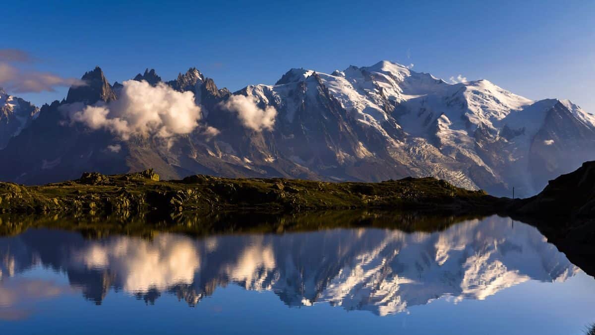 A range of mountains against a blue sky, reflected in a lake