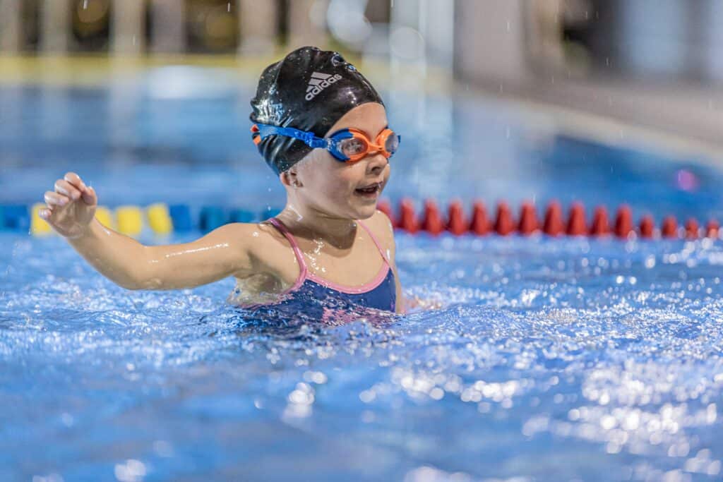 A young girl wearing a swimming cap and goggles swims in an indoor pool