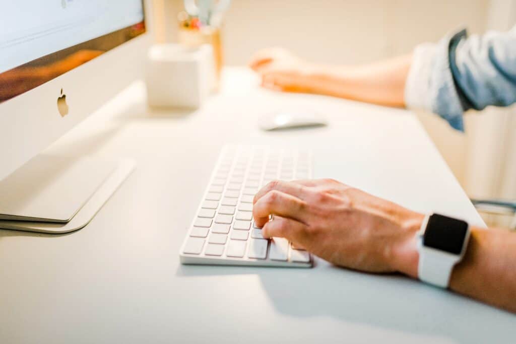 La main d'un homme au premier plan posée sur un clavier blanc en face d'un écran d'ordinateur. 