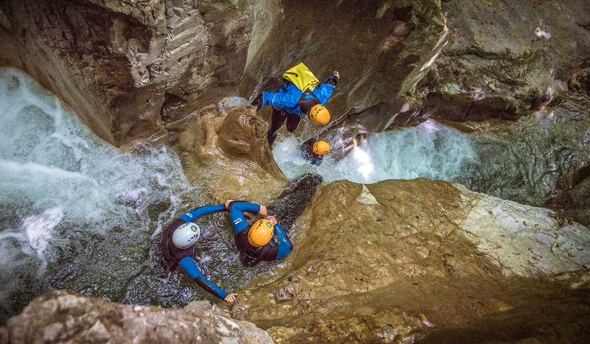 Four people wearing wetsuits and helmets slide down a mountain stream