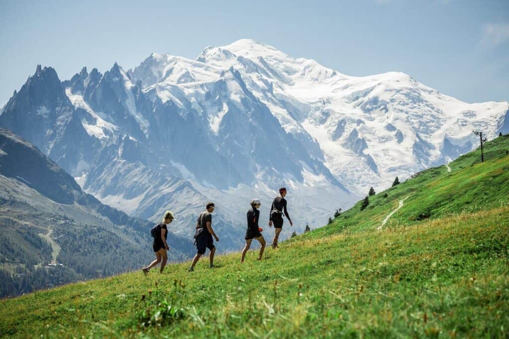 Four walkers head up a grassy mountain slope, with snow-covered peaks in the background